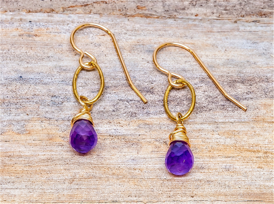 Pair of gold dangle earrings with faceted teardrop-shaped purple gemstones wrapped in gold wire, displayed on a rustic wooden background