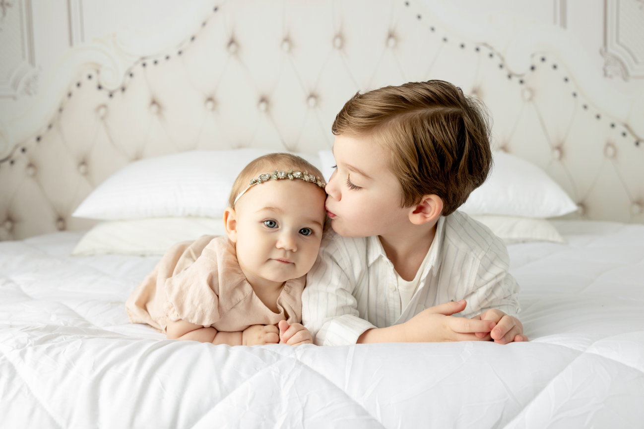A young child kisses a baby's head on a white bed with tufted headboard in the background.