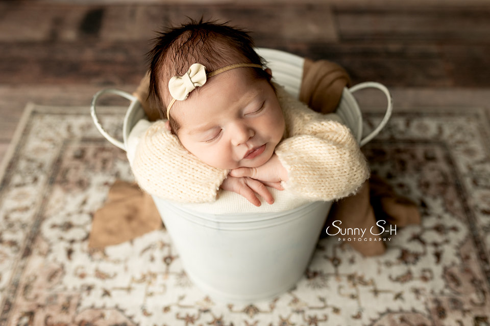 Sleeping baby in a white bucket, wearing a headband, on a textured rug with wooden flooring.