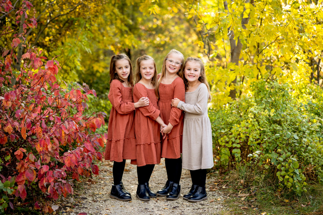 Four young girls in autumn dresses stand on a leafy path, surrounded by vibrant fall foliage.