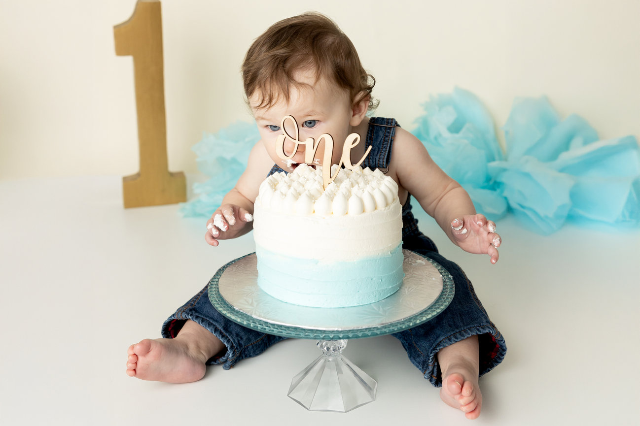 Baby in denim outfit sits near blue and white cake with one topper, large number one prop in background.