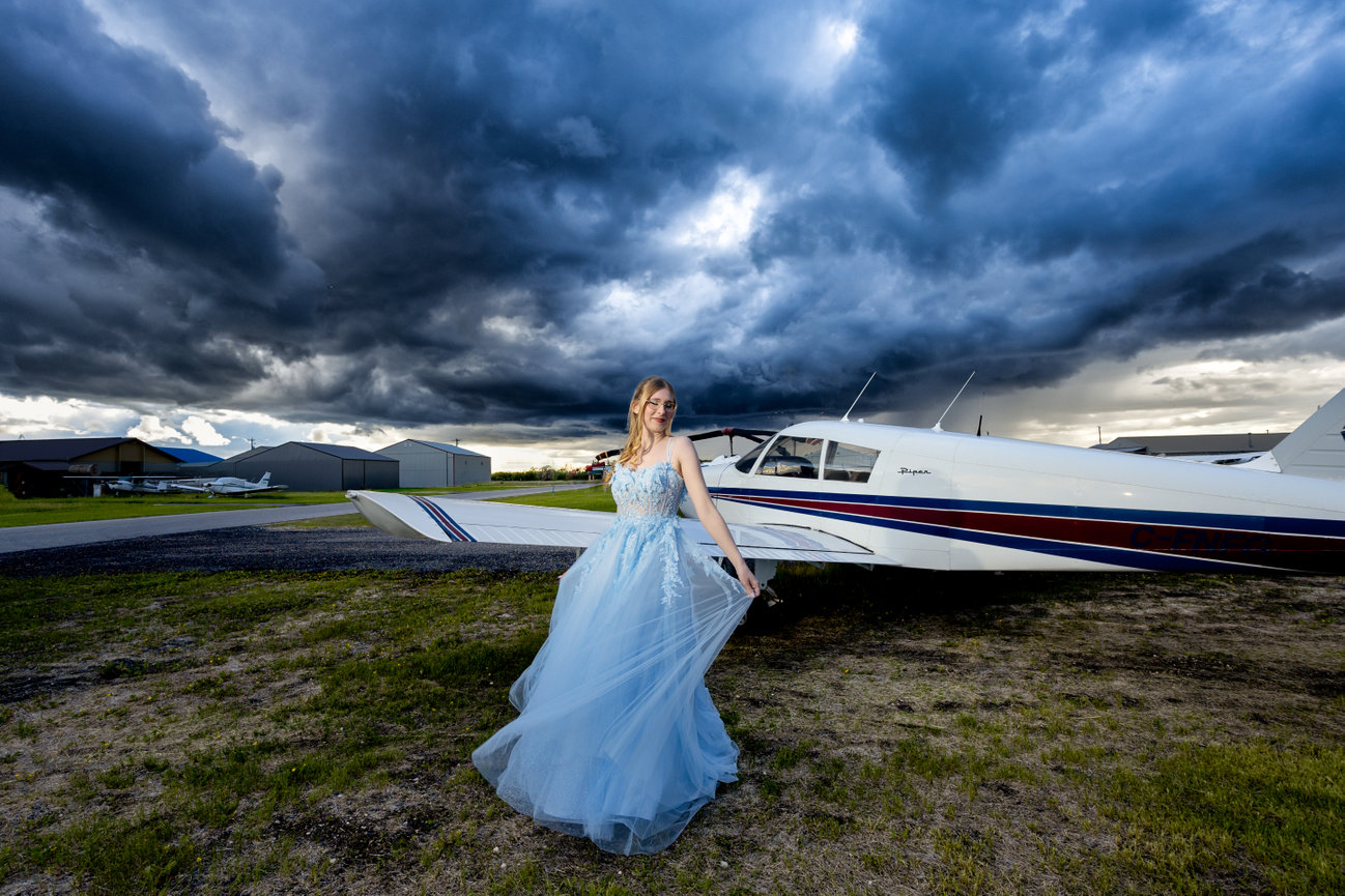Graduate in a blue gown posing beside a small airplane under dramatic storm clouds