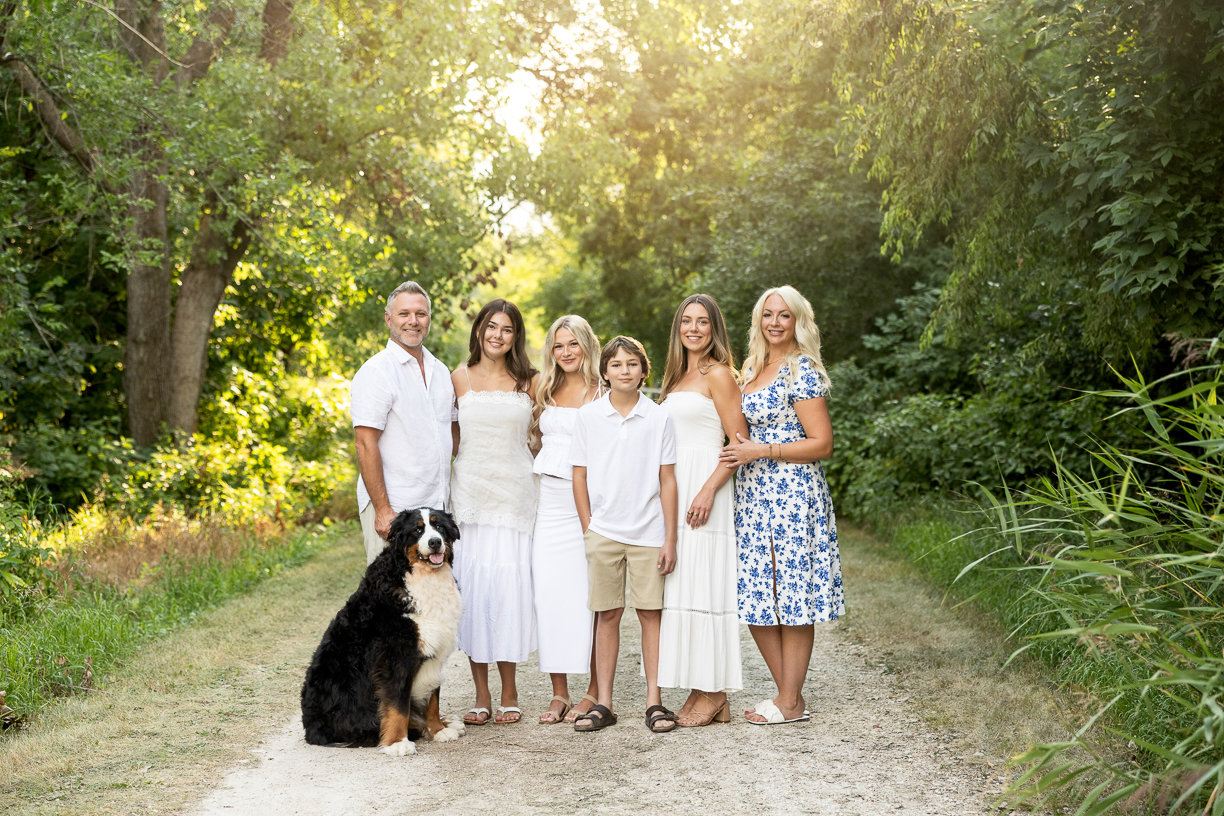 Family of six and a dog posed in white clothes with summer tree and sun background in winnipeg
