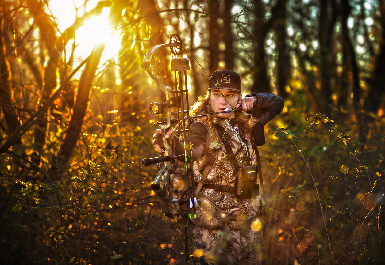 Person aiming a bow in a golden forest at sunset.