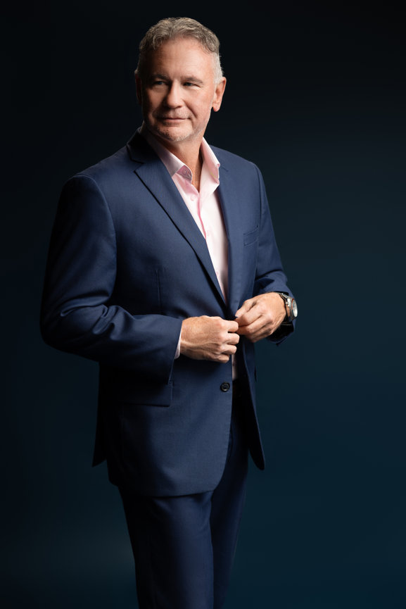 Professional branding headshot of a businessman in a navy suit smiling confidently against a dark blue backdrop.