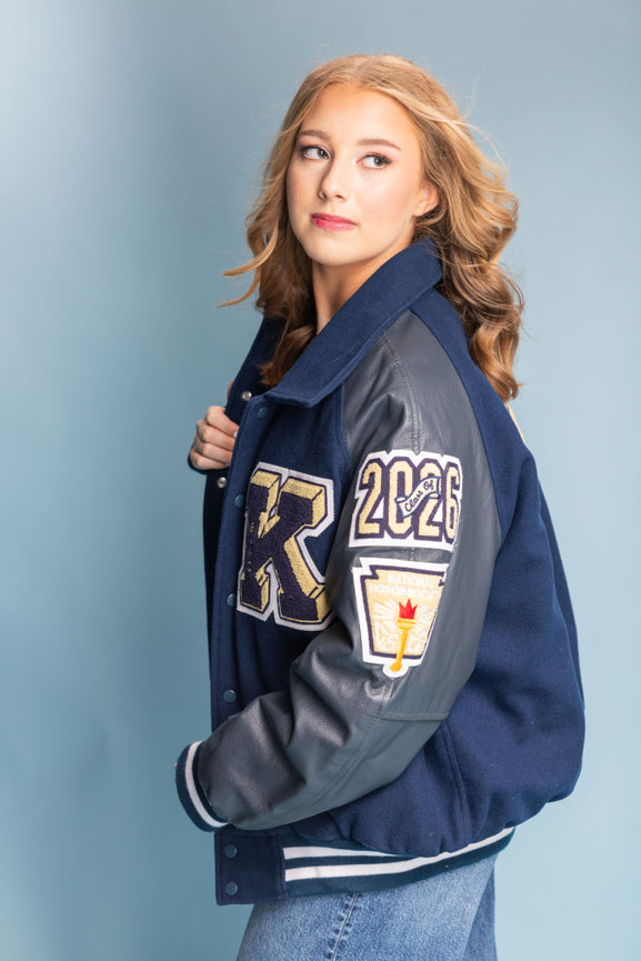 A person in a navy letterman jacket with patches, against a light blue background. high school senior photo