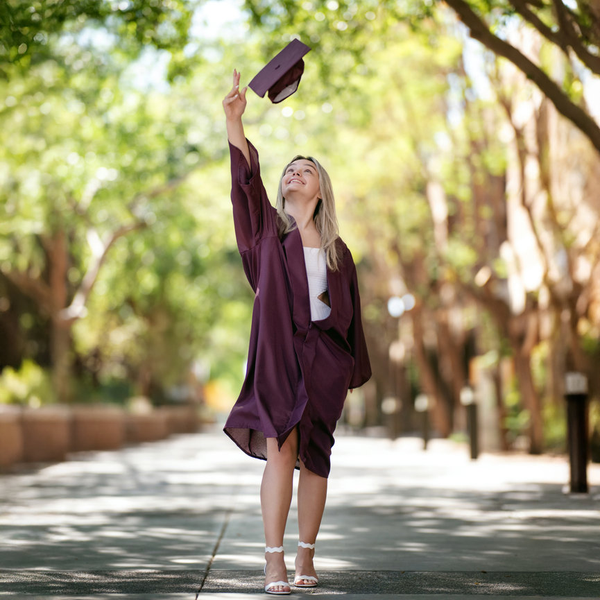 Senior girl in maroon graduation gown walking down tree-lined path ASU West