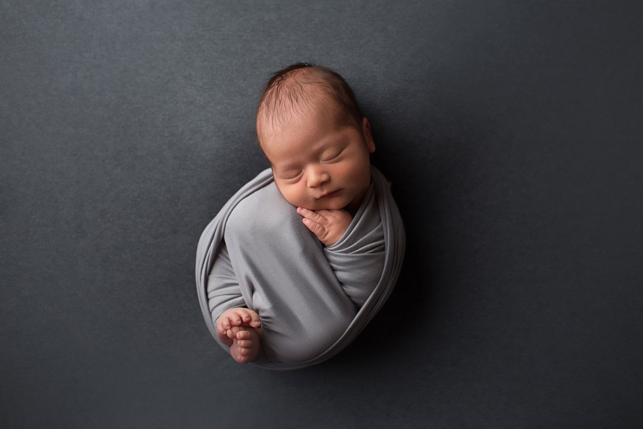 Newborn baby boy curled up sleeping in light gray wrap on dark gray background at newborn photo session with Kansas City photographer Dreamworks Photography