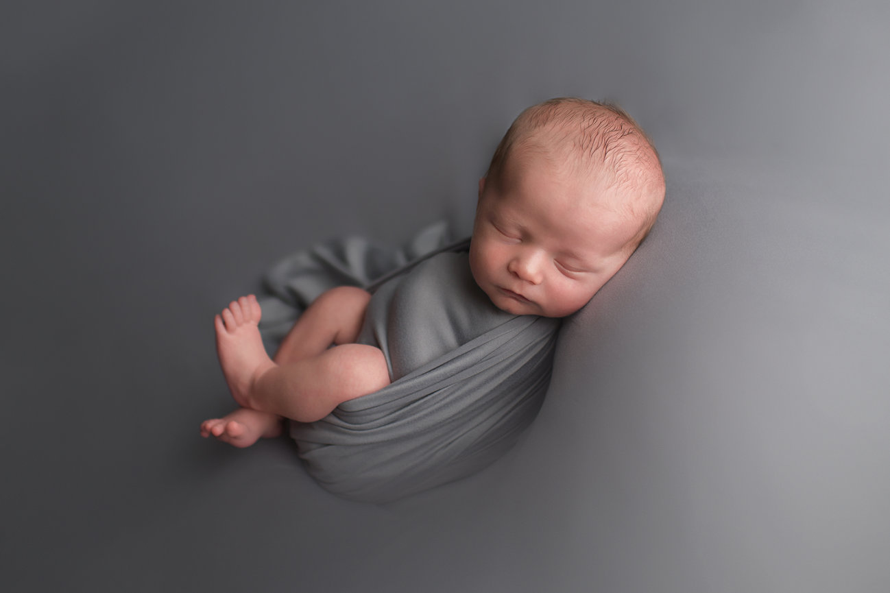 Newborn baby sleeping on back curled up in a grey wrap on a grey backdrop with Kansas City newborn photographer Dreamworks Photography