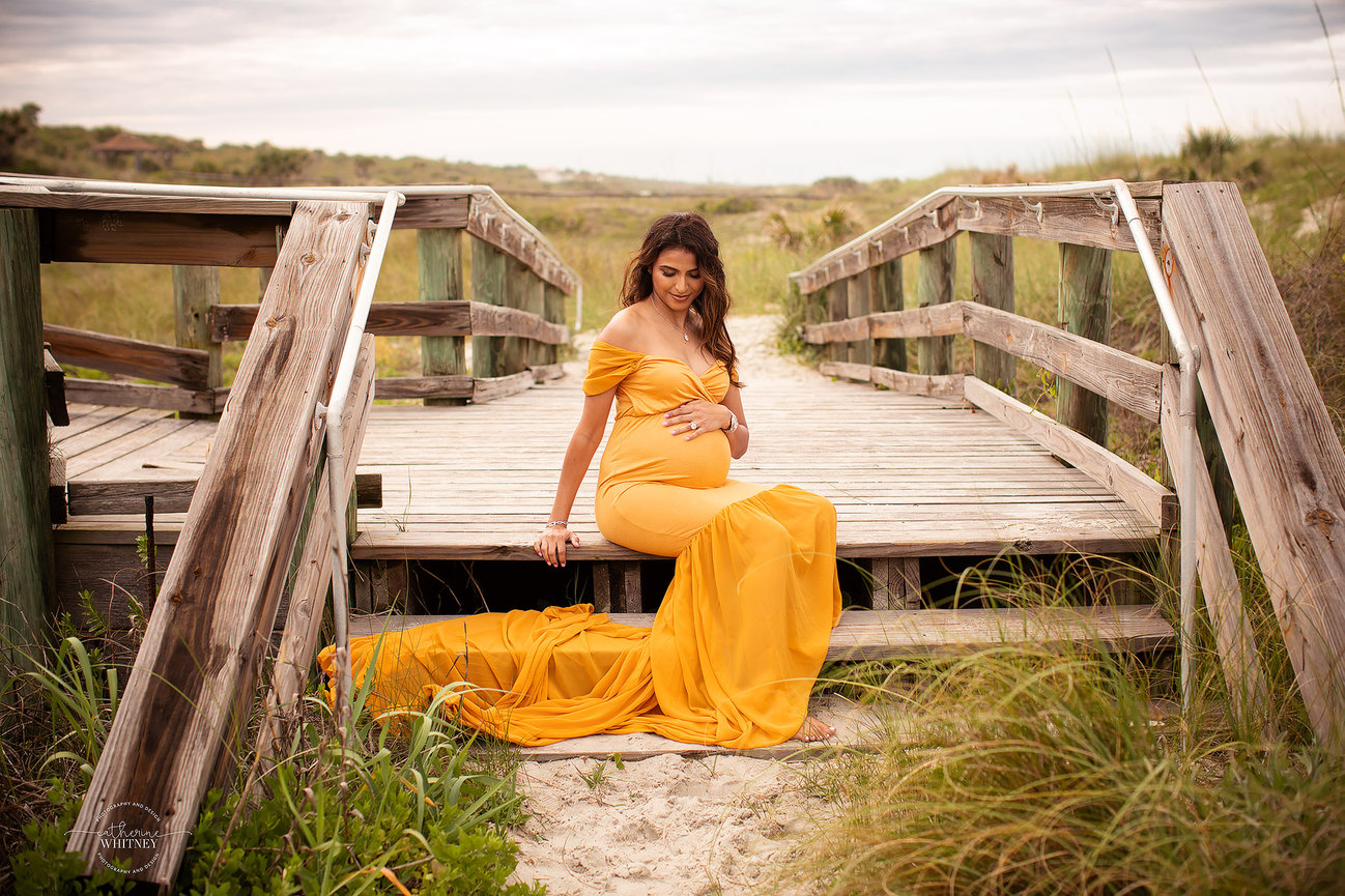 Pregnant woman in a flowing yellow dress sits on a wooden walkway, surrounded by grass and a cloudy sky at Hanna Park Beach during a maternity photography session with Catherine Whitney Photography in Jacksonville Florida