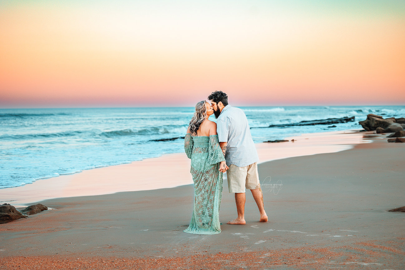 Couple walking hand in hand on a beach at sunset, with waves and a colorful sky in the background in St. Augustine during a maternity photography session with Catherine Whitney Photography in Florida