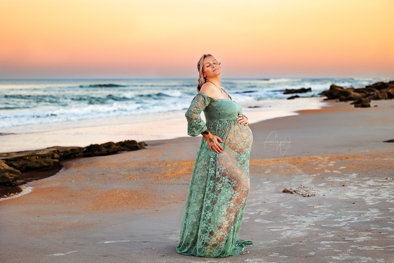 Pregnant woman in a green lace dress stands on a beach at sunset, with waves in the background during a maternity photography session with Catherine Whitney Photography in St. Augustine Florida