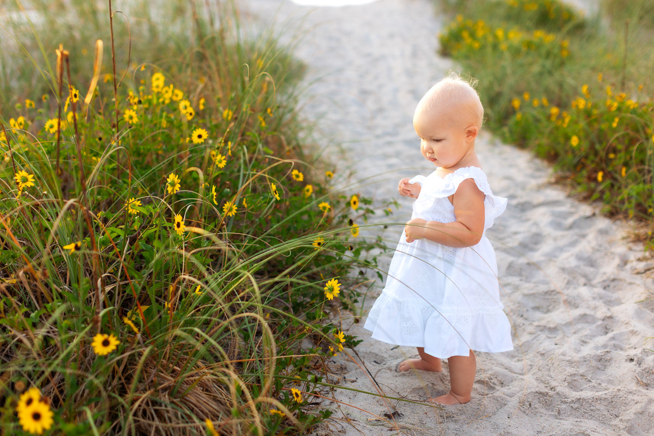 A baby wearing a white dress stands on a sandy path to the beach at Anastasia Island, Florida, enjoying the warm sun and soft sand beneath their feet, smelling flowers during a family photography session with Catherine Whitney Photography