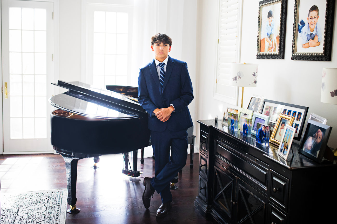 A high school senior in formal attire stands by a piano, exuding confidence and charm during his senior photography session with Catherine Whitney Photography