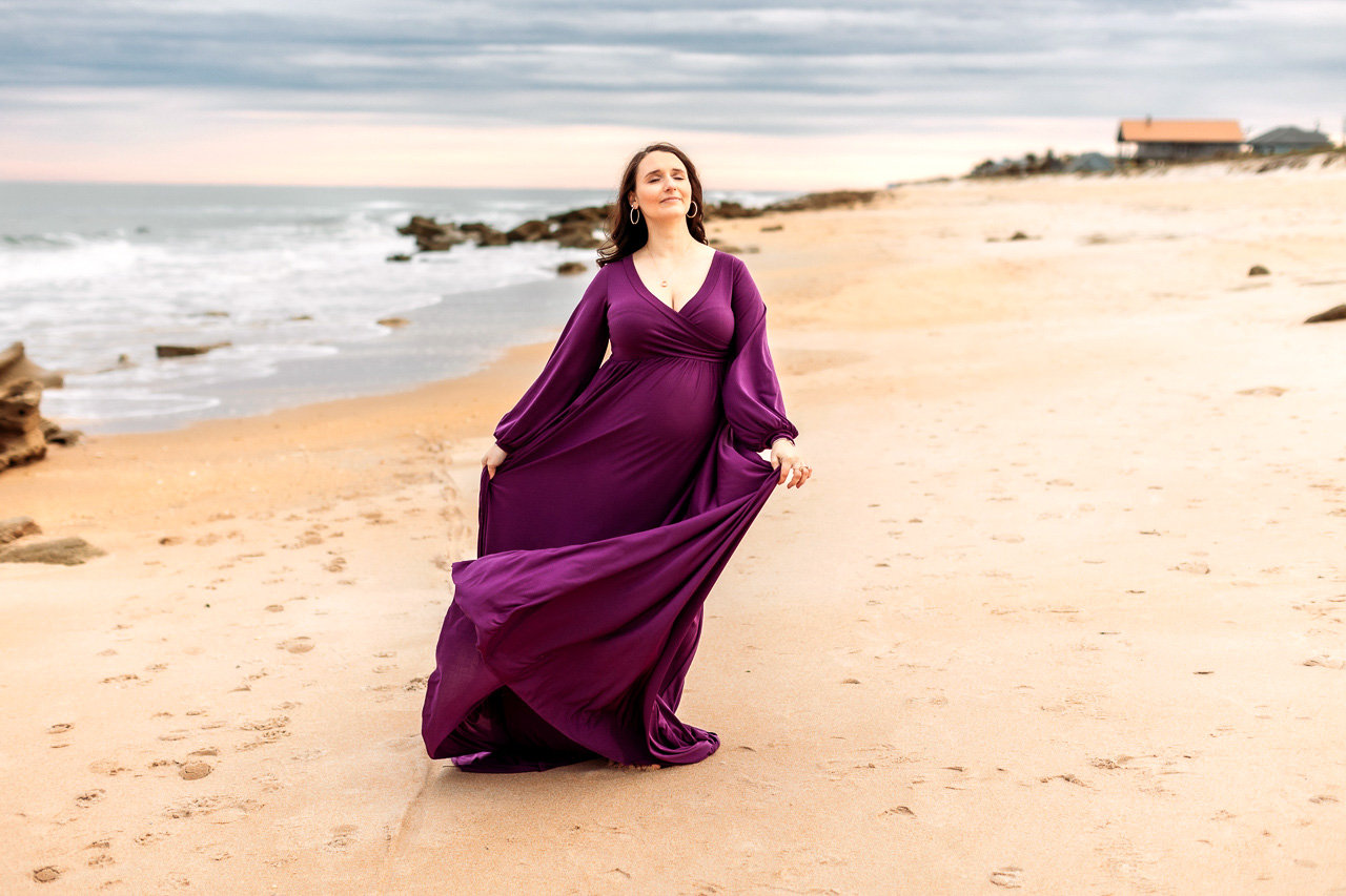 Woman in a flowing purple dress walking on a sandy beach with waves and cloudy sky in the background during a maternity photography session with Catherine Whitney Photography in St. Augustine Florida