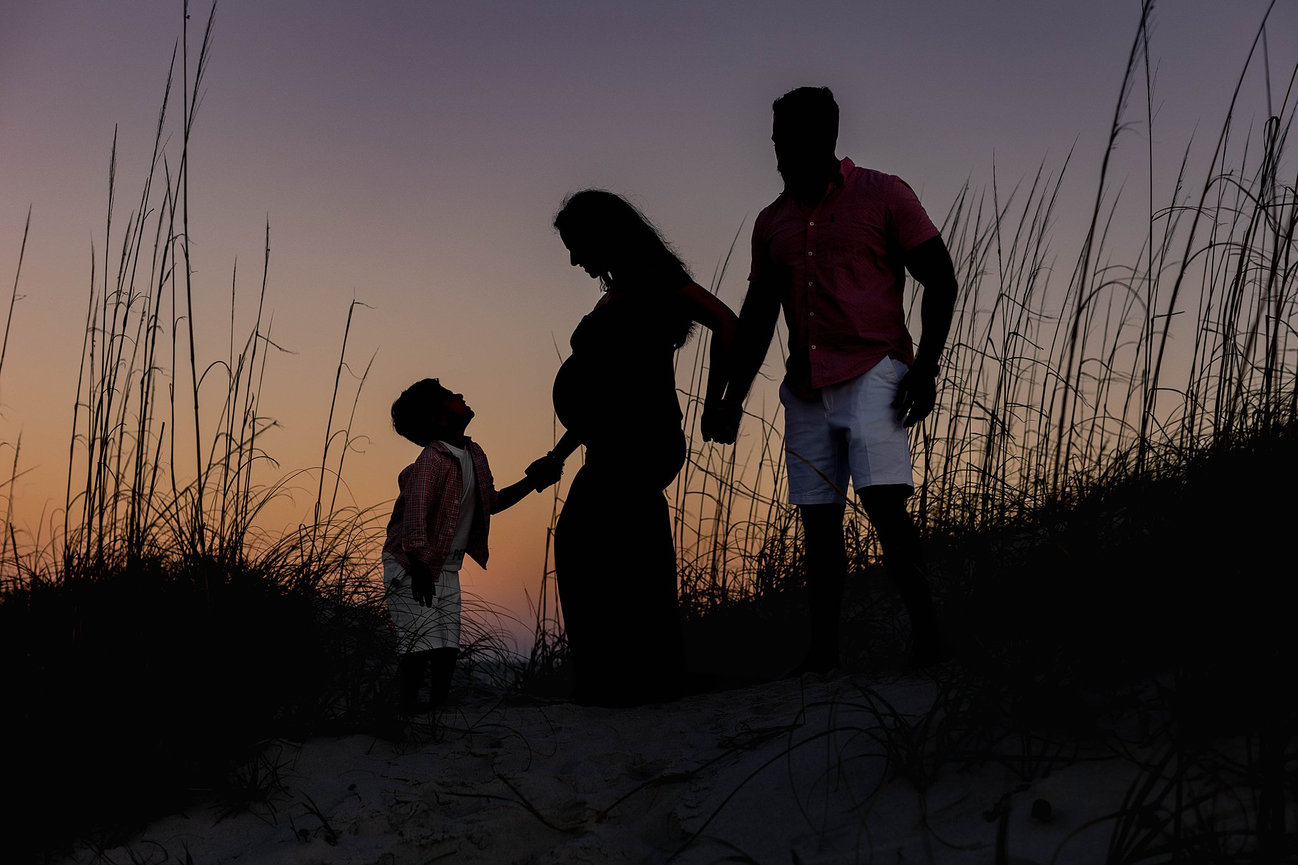 Silhouette of a family on a beach at sunset; a pregnant woman, a man, and a child hold hands at Hanna Park Beach during a maternity photography session with Catherine Whitney Photography in Jacksonville Florida