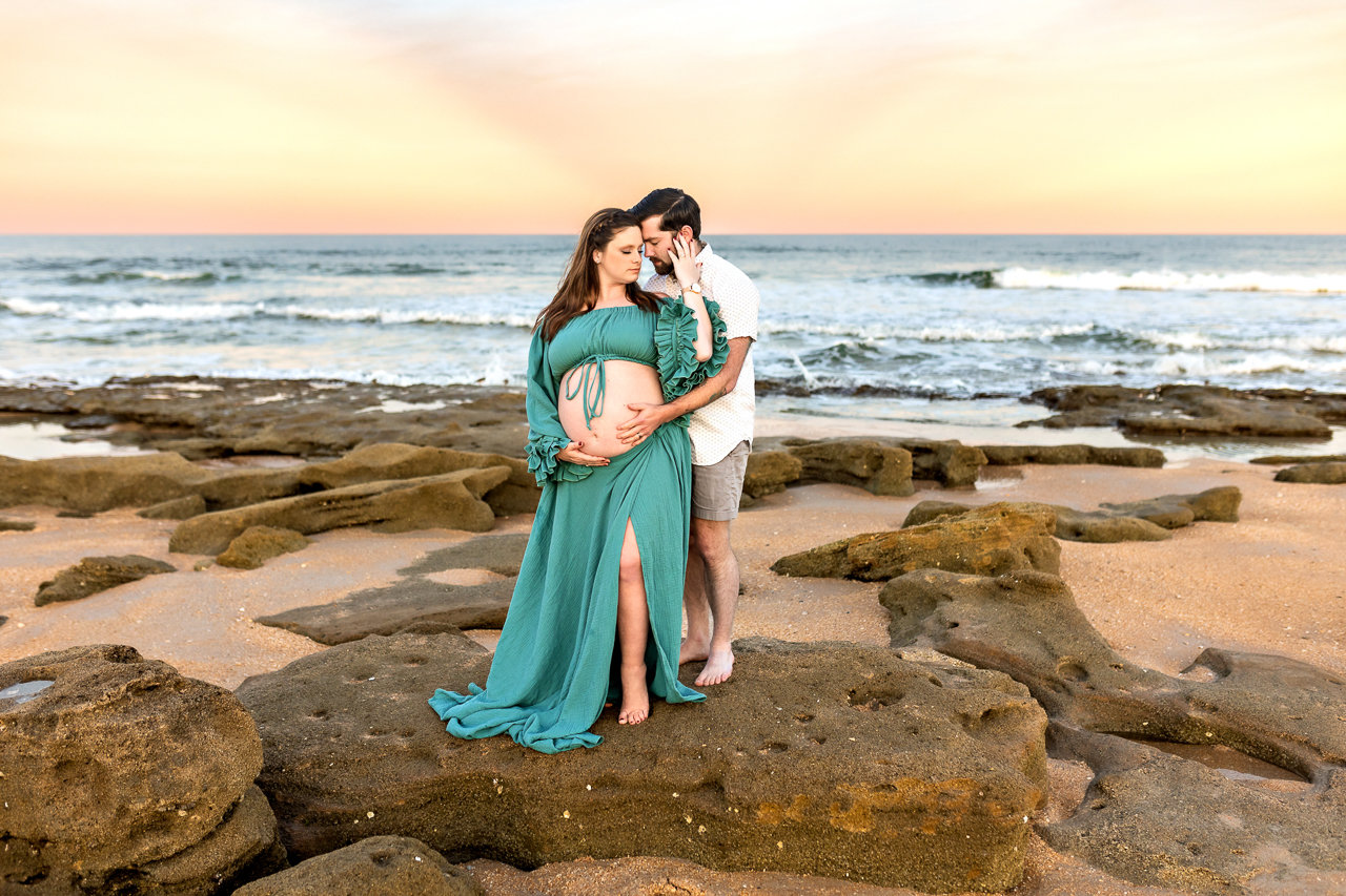 Couple embracing on rocky beach at sunset with ocean in background in St. Augustine during a maternity photography session with Catherine Whitney Photography