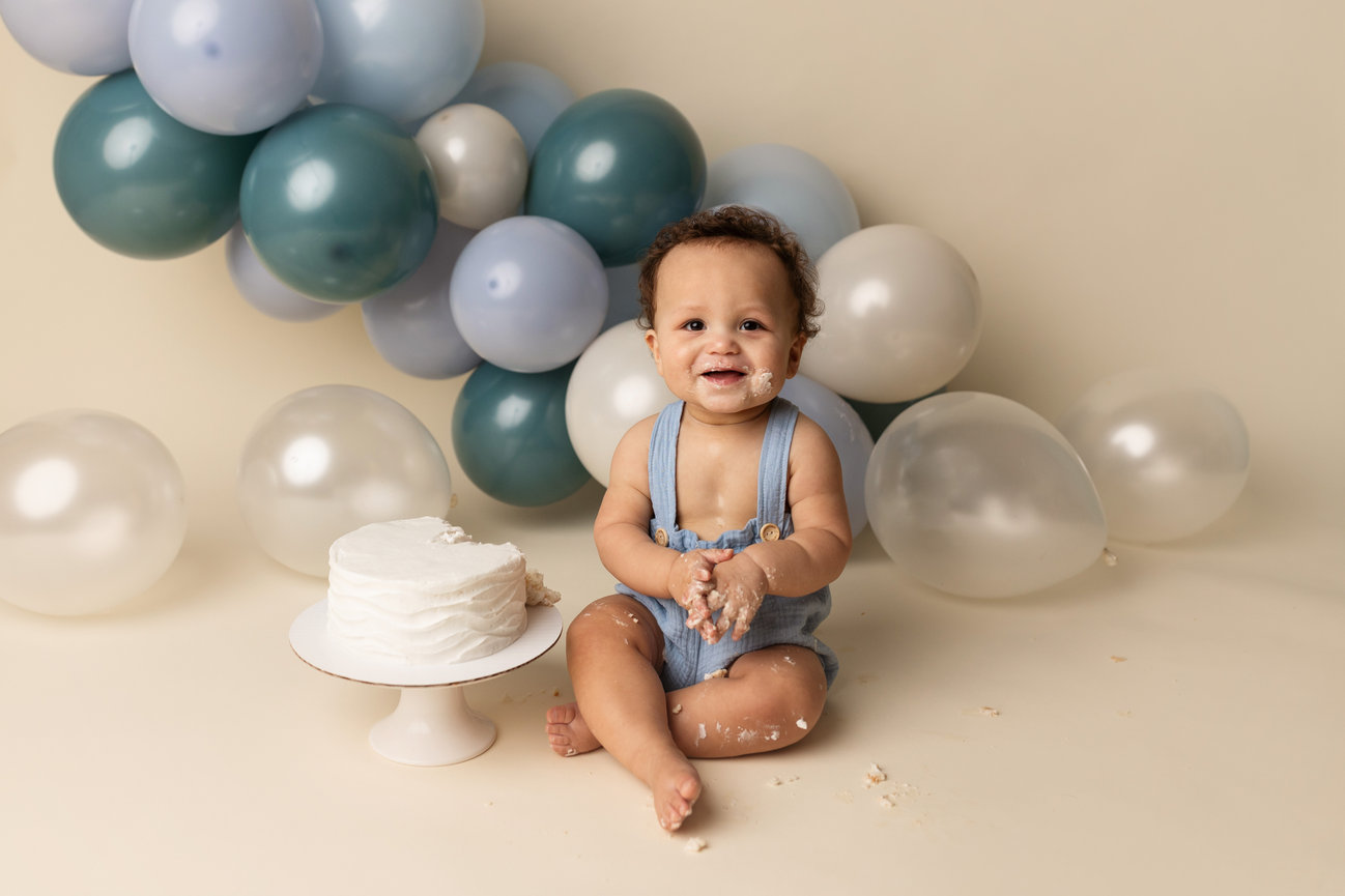 Omaha Cake Smash Photographer with cake on their face sits near a cake, surrounded by blue and white balloons.
