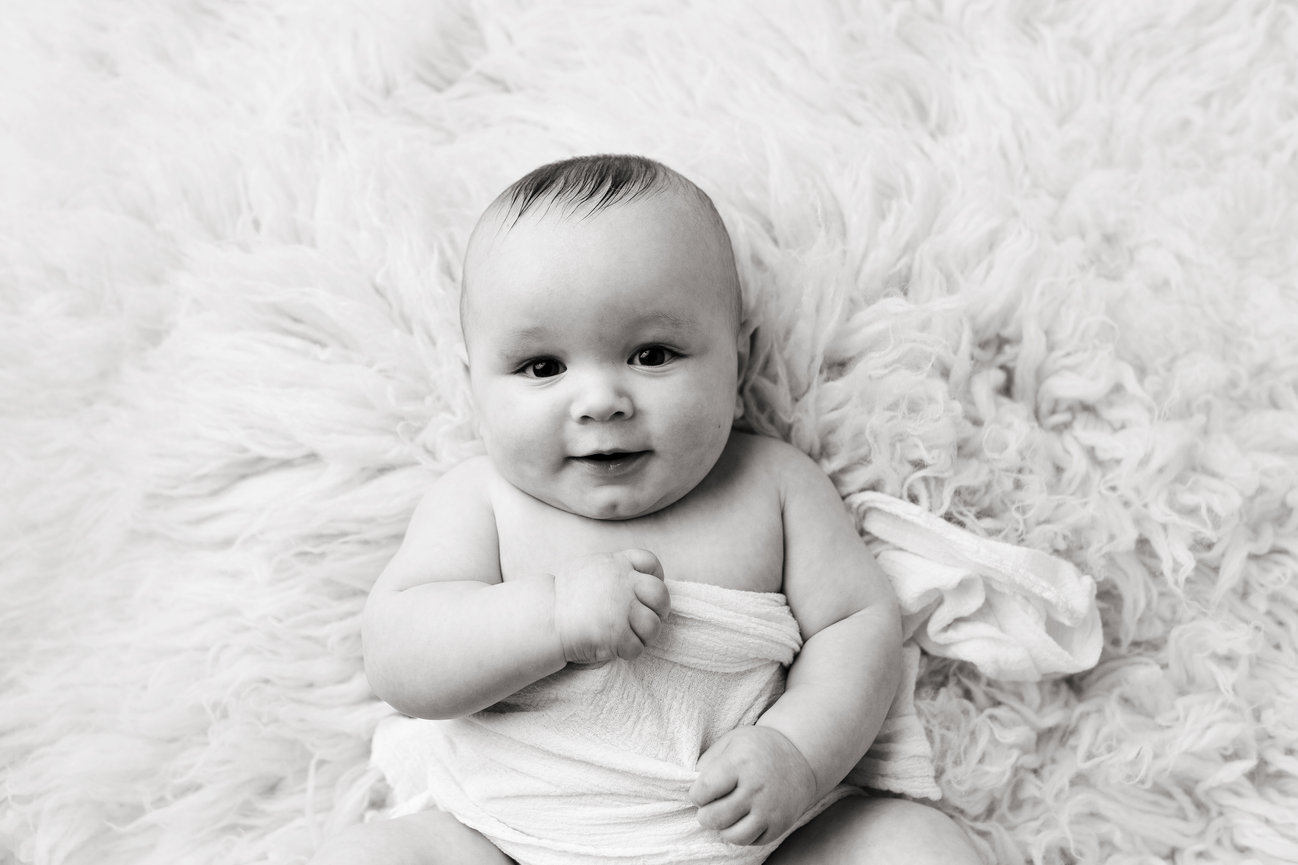 Omaha Baby Photographer captured a smiling baby wrapped in a blanket lying on a fluffy surface, black and white.
