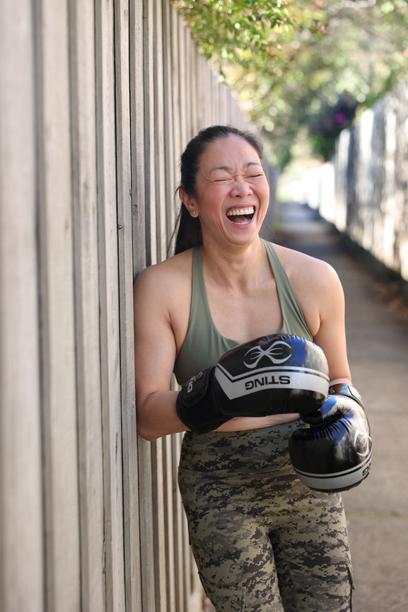 Laughing woman in boxing gloves and workout gear leaning against a wooden fence outdoors. authentic photos for my business and website