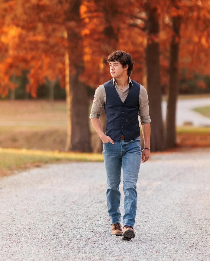 A high school senior strolls on a gravel path, wearing jeans and a navy vest over a shirt. The scene is set in autumn, with trees adorned in vibrant orange leaves surrounding the path.