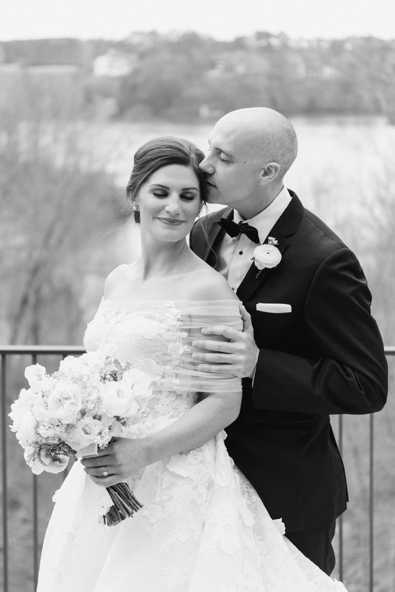 Black and white image of groom kissing bride on cheek by the lake. Wedding photography by Heather Durham.