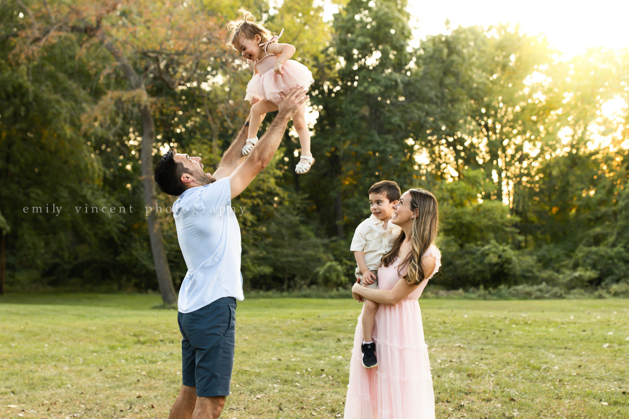 Playful moment of family watching dad throw little girl in the air in grassy field