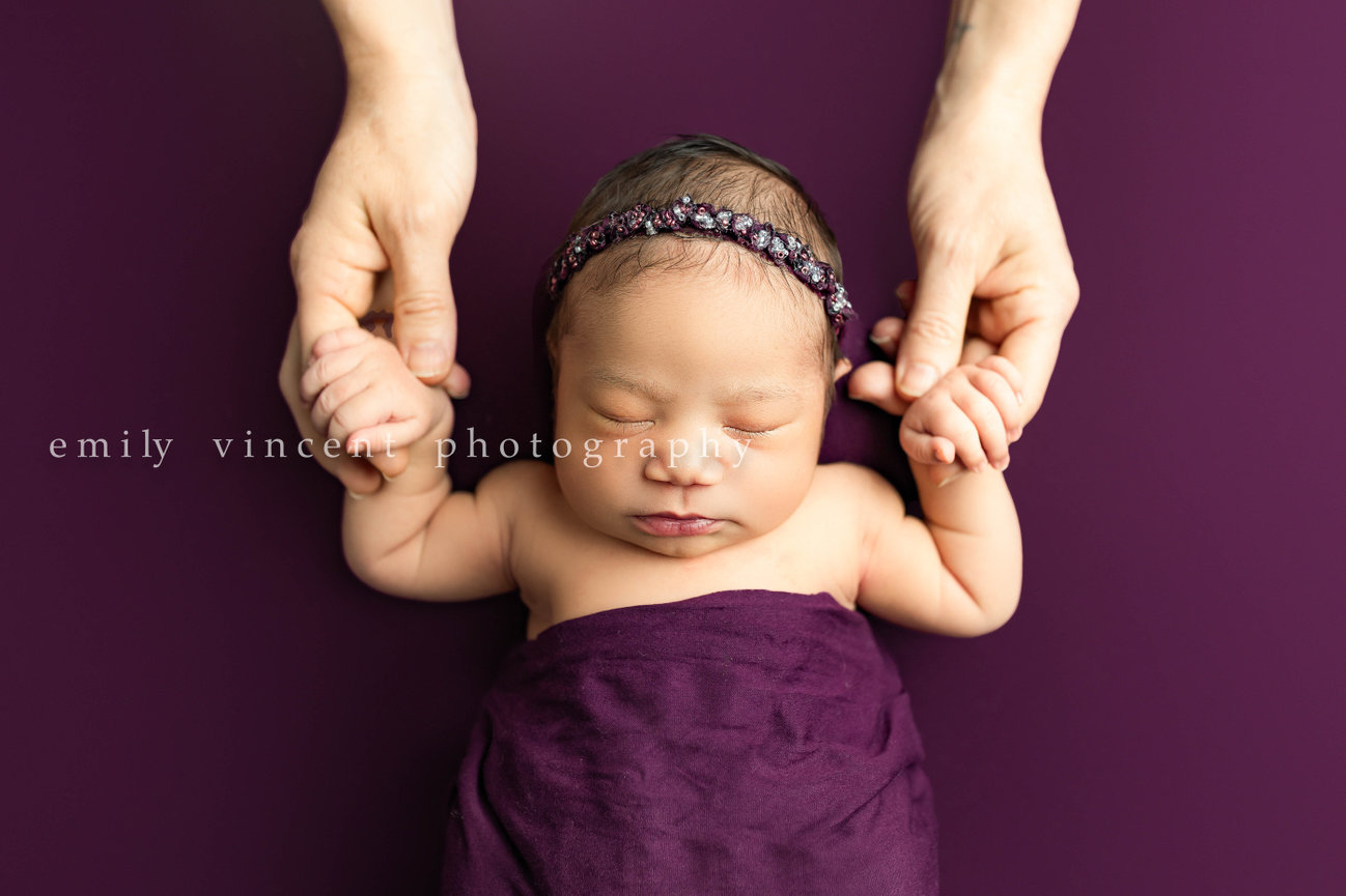 Newborn baby with headband in purple blanket with mother holding hands