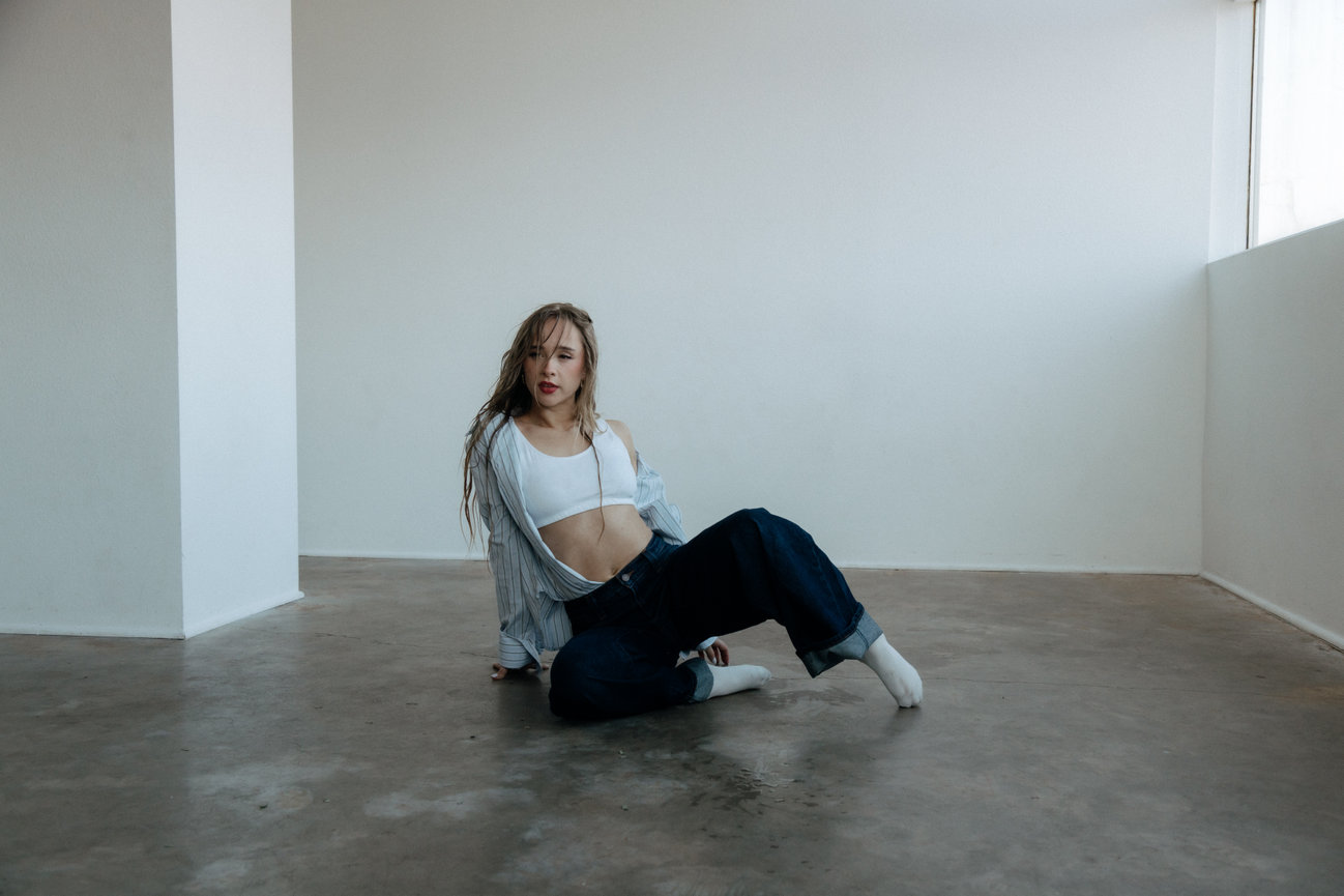 Person sitting on a concrete floor in a minimalist room wearing a crop top, jeans, and a striped shirt.