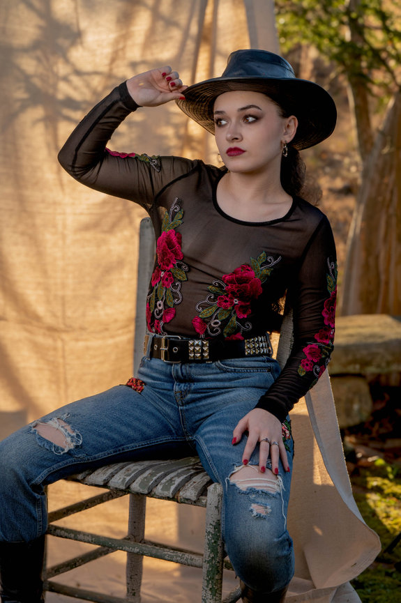 A woman in a sheer top with floral designs, ripped jeans, and a wide-brimmed hat sits on a rustic chair outdoors captured by Jo Hayes Images Charlotte Event Portrait Fashion Photographer