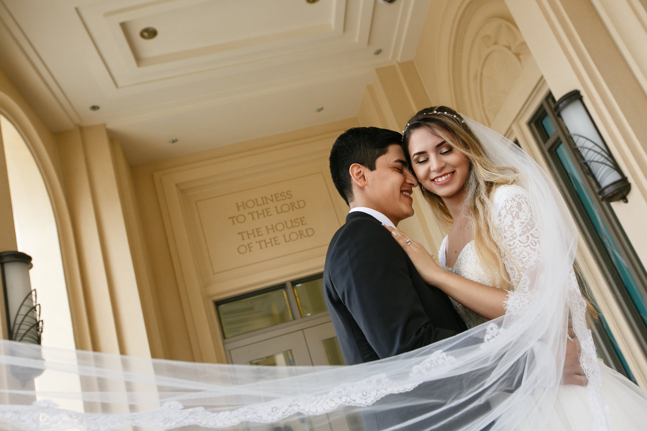 A bride and groom embrace under a building archway, with the bride's veil flowing.