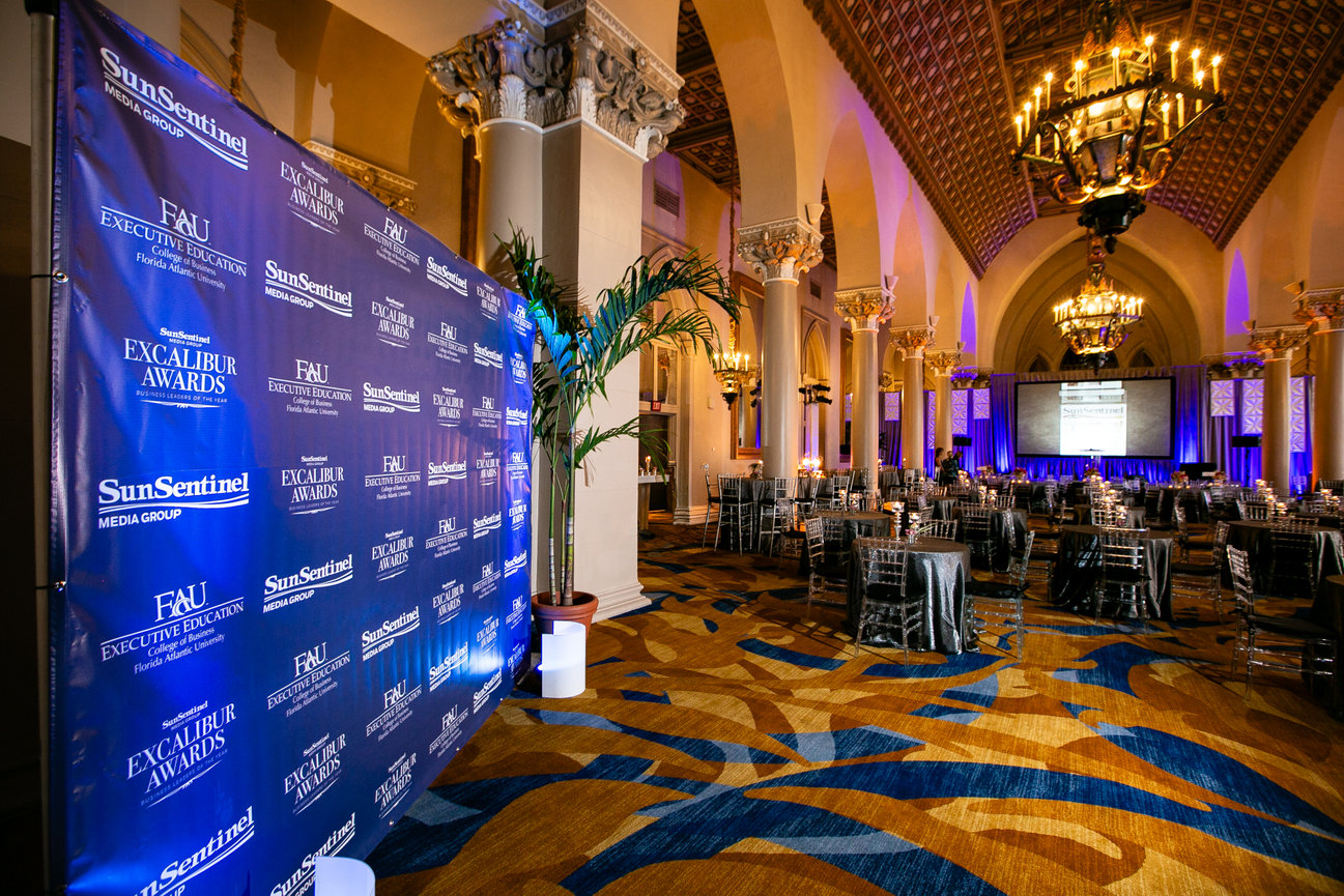 Elegant awards banquet in a grand hall with chandeliers, round tables, and a SunSentinel Excalibur Awards backdrop