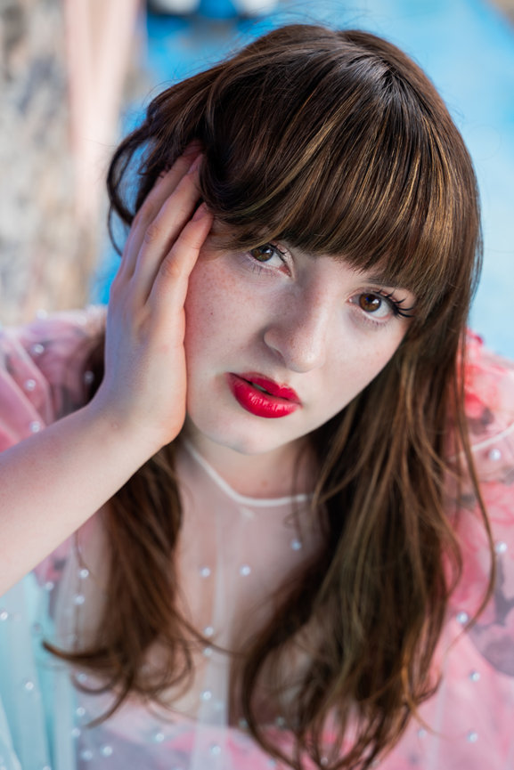 Close-up portrait of a young woman with brown hair, red lipstick, and hand on her face against a blue background