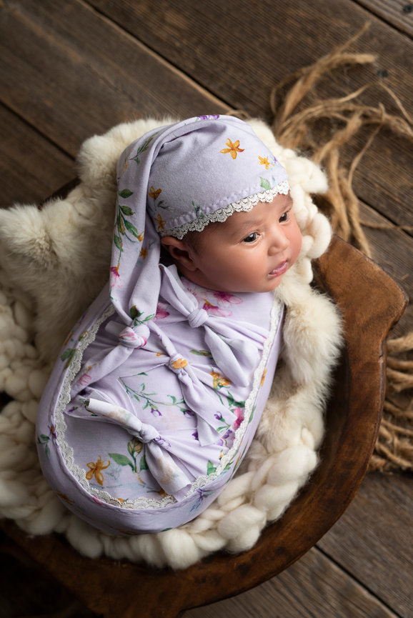 A baby is swaddled in a purple, floral-patterned blanket and matching hat, lying on a fluffy, cream-colored rug in a wooden bowl. The baby gazes upward with wide eyes, set against the rustic charm of a Barrington, Illinois wooden floor.