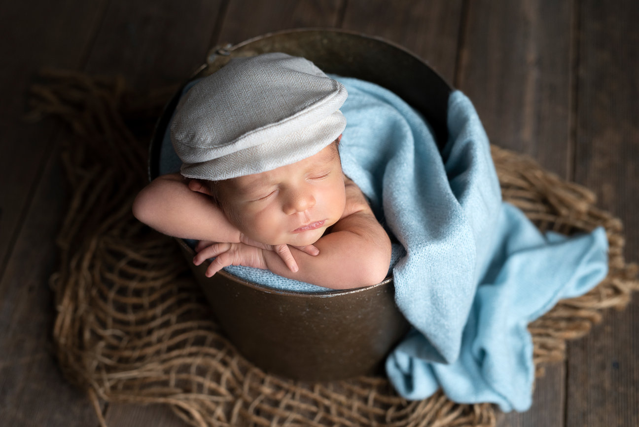In a serene Lake in the Hills studio, a newborn baby peacefully sleeps in a small metal bucket. Wrapped in a soft blue blanket and wearing a white cap, the little one rests on a wooden floor with a burlap mat underneath.