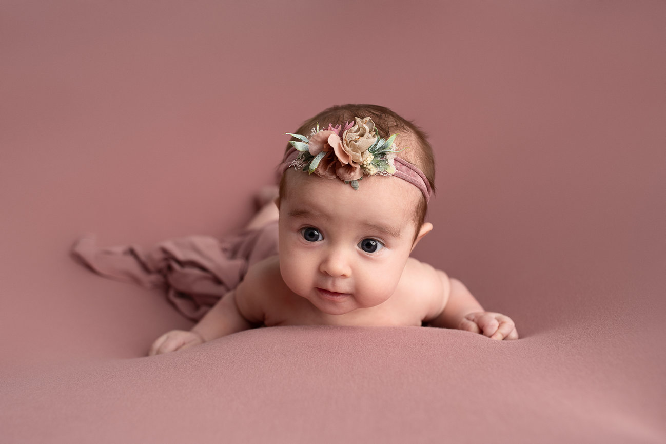 A baby lying on their stomach against a soft, pink backdrop, reminiscent of a Chicago sunrise. They wear a floral headband adorned with pastel blooms. With wide eyes, they look curiously at the camera, found in the heart of Chicago.