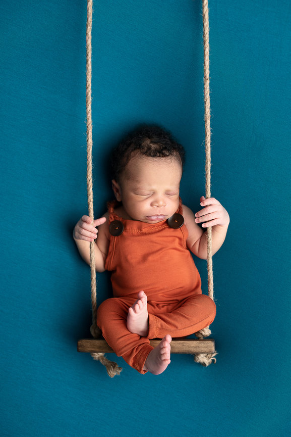 A baby in an orange outfit is peacefully asleep on a small wooden swing, reminiscent of the serene landscapes of Algonquin. The swing, suspended by rope against a teal background, cradles the baby whose eyes are closed and legs are curled up gently.