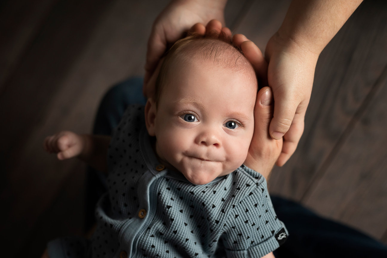 A baby with expressive eyes lies on a dark surface, wearing a light gray dotted outfit. The little one has a small, thoughtful expression, gently cradled by an adults hands. This precious moment highlights the importance of baby care classes near me.
