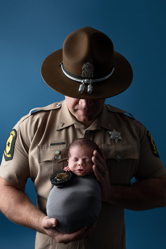 A uniformed officer from Gilberts, Illinois, gently cradles a sleeping newborn wrapped in a gray blanket. The officer wears a brown hat and beige shirt adorned with badges. The baby restsin the officers hands, against a soft blue background.