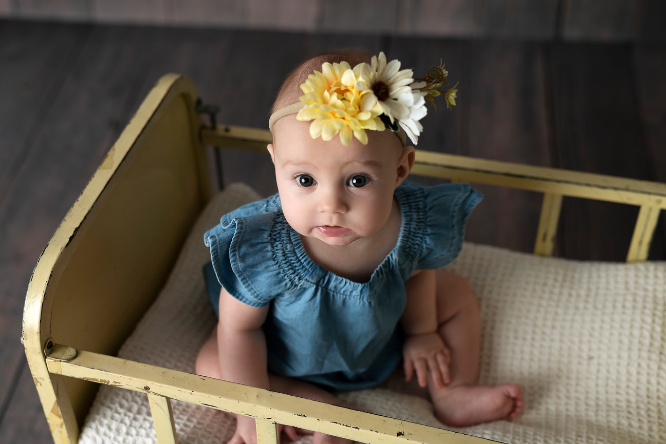 A baby, perfect for your next photoshoot idea, wears a blue dress and flower headband as she sits on a small vintage bed with a white quilt. The dark wooden floors in the background. Consider this charming setup among places to take pictures near me.
