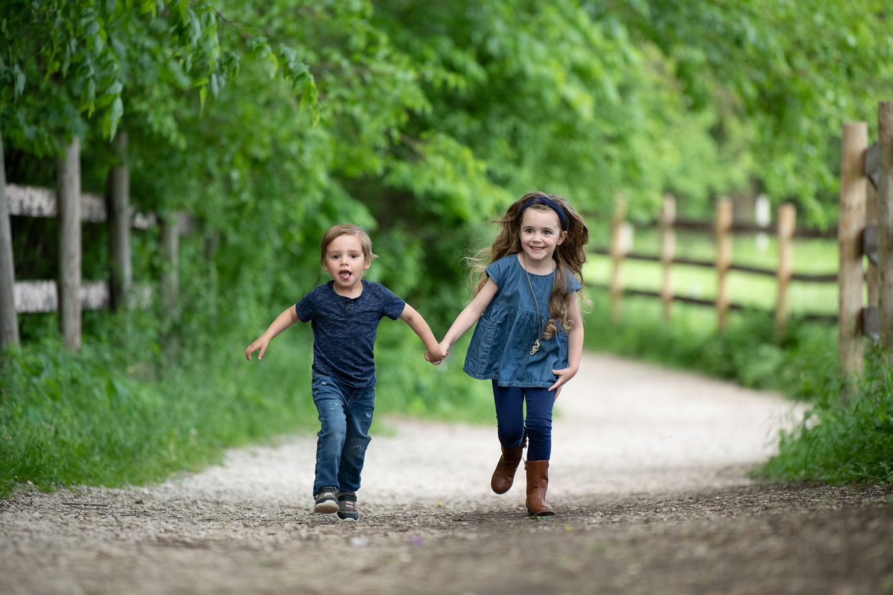 Two young children hold hands and run happily down a gravel path surrounded by lush green foliage and wooden fences in a perfect setting for North Barrington family photography. Both wear blue shirts and jeans, with one sporting brown boots.