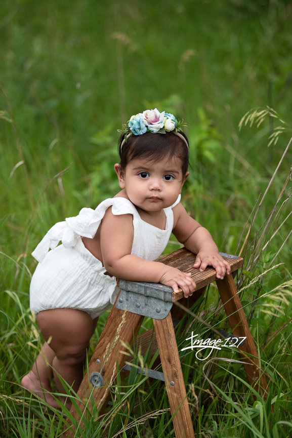 A baby wearing a white outfit and floral headband stands against a small wooden ladder on the grassy terrain of Crystal Lake, Illinois, looking towards the camera.