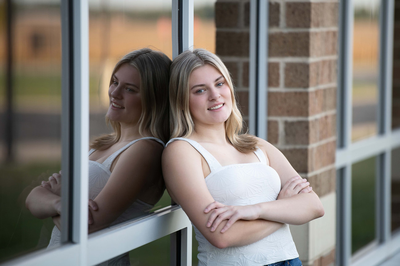 A young woman with blonde hair stands outside, leaning against a brick wall with her arms crossed. Smiling, her reflection is captured in the window beside her. The scene, reminiscent of high school photos, has a blurred view of grass background.