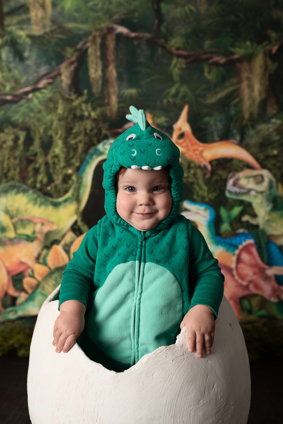 A talented photographer captures a baby in a green dinosaur costume nestled inside a large white eggshell. The backdrop is alive with colorful illustrations of dinosaurs and a lush, forested setting.