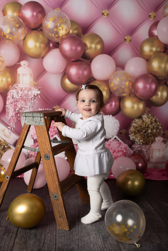 beside a wooden step ladder, celebrating her birthday. The background is adorned with pink and gold balloons and decorations, creating a festive atmosphere. Some balloons are also scattered on the dark wooden floor.