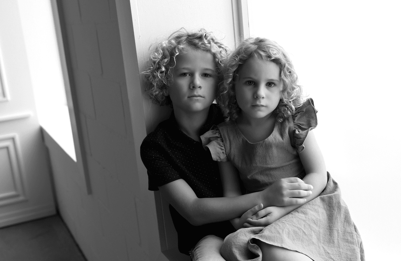 A four year old sister rests in the arms of her older brother in the window sill in the Kliks Photography studio.