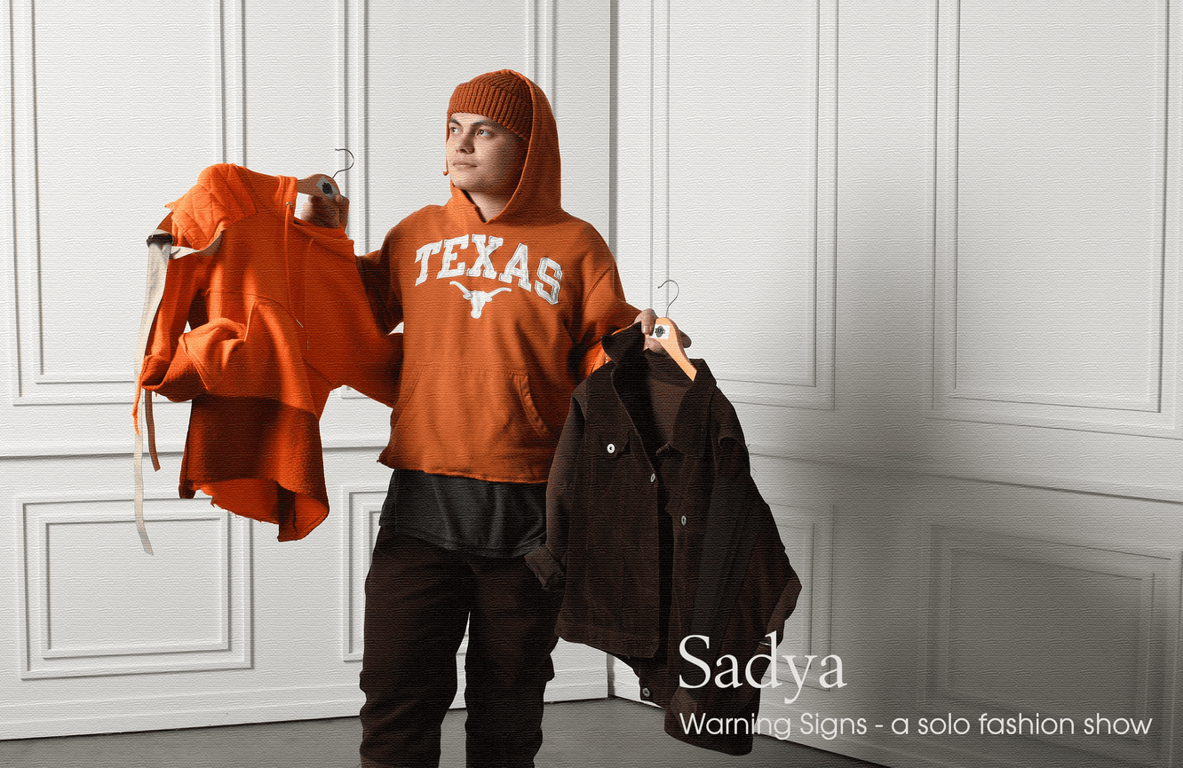 A male designer in an orange hoodie holds fashion pieces that he designed.