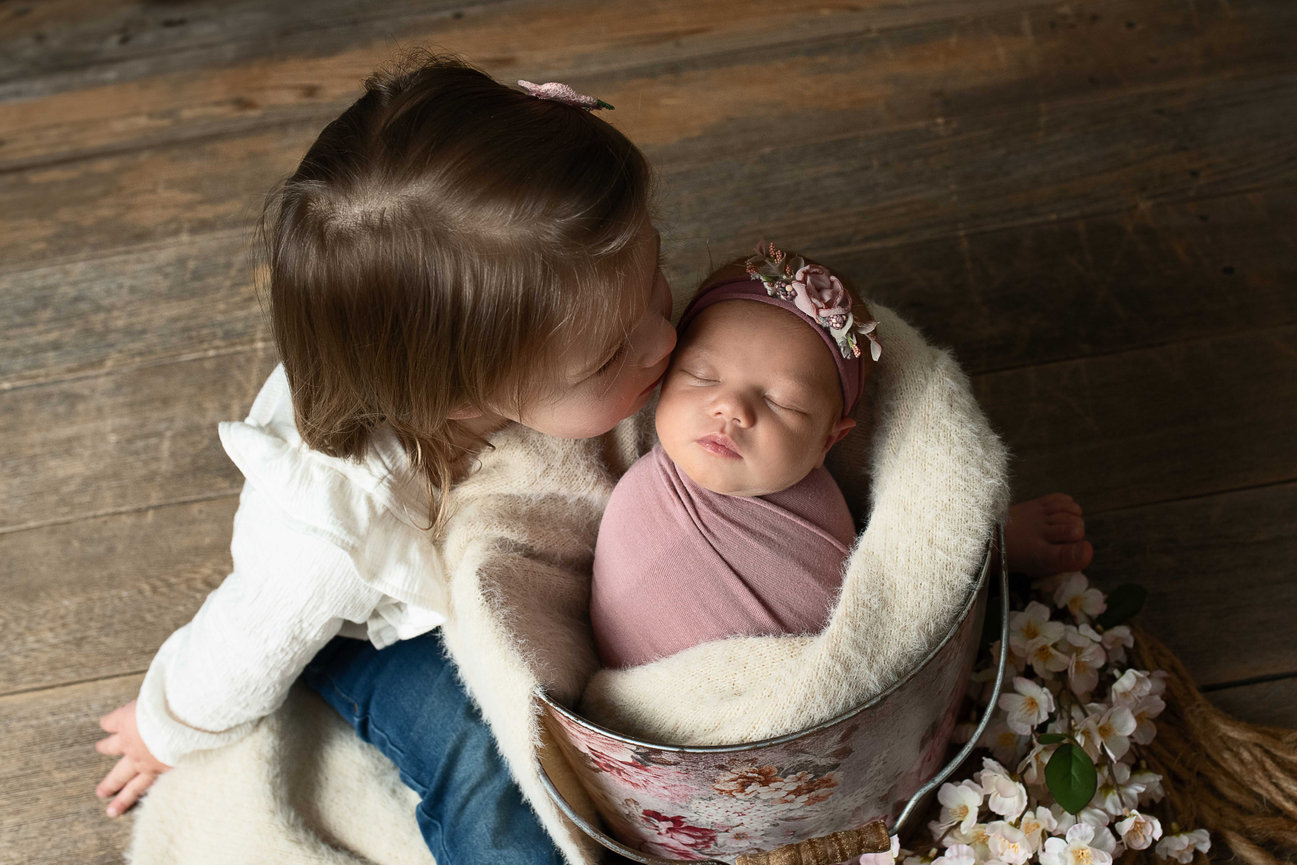 Big sister gently kisses newborn baby girl swaddled in pink with floral headband, posed in a floral bucket – sibling newborn photography in Crystal Lake, Illinois