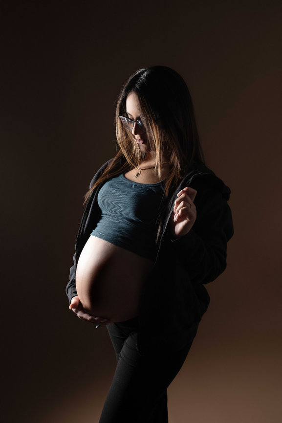 Pregnant woman in a dark setting, wearing sunglasses, holding her belly and looking down, with a gentle light focus.