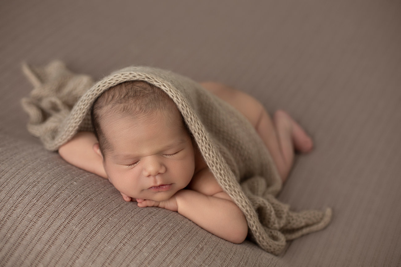 Newborn boy posed with his hands under his chin. He is lying on a beige backdrop and a knitted wrap is draped over him.
